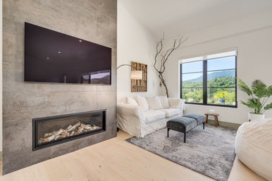 Bright modern living room with wall-mounted TV and linear fireplace, white slipcovered sofa, gray ottoman, patterned rug, potted plant, and large window showcasing green hillside and mountain views.
