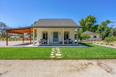 Modern farmhouse-style cottage with covered front porch and rocking chairs, stone stepping path across green lawn, gravel driveway, wooden carport and outbuilding under a clear blue sky.