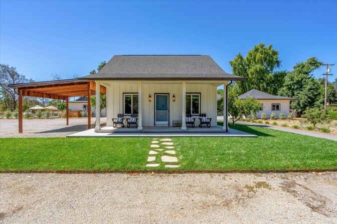 Modern farmhouse-style cottage with covered front porch and rocking chairs, stone stepping path across green lawn, gravel driveway, wooden carport and outbuilding under a clear blue sky.
