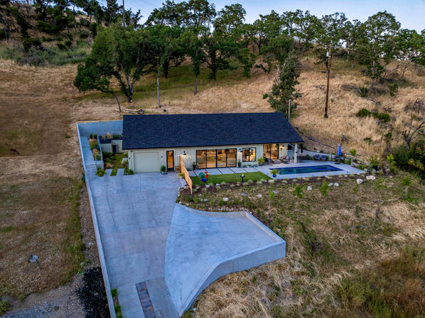 Aerial view of a modern single-story ranch home on a golden hillside, with a long concrete driveway and attached garage, glass patio doors opening to a rectangular swimming pool and lawn, surrounded by oak trees and dry grassland.