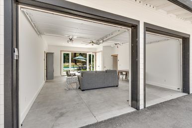 Open two-car garage converted into a bright living area with concrete floors, gray sofa, ceiling fans, and sliding glass doors opening to a backyard patio and pool with umbrella.