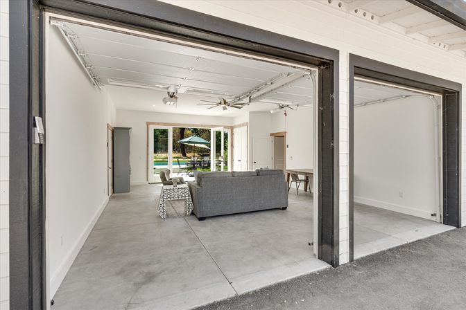 Open two-car garage converted into a bright living area with concrete floors, gray sofa, ceiling fans, and sliding glass doors opening to a backyard patio and pool with umbrella.
