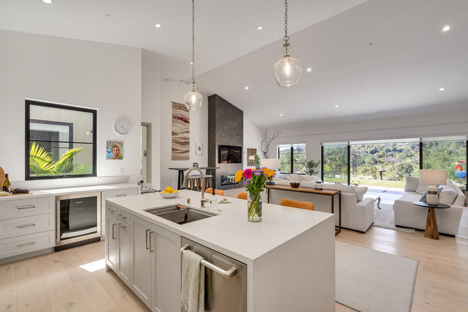 Bright modern open-concept kitchen and living room with white island and sink, glass pendant lights, colorful flower vase, and sliding glass doors opening to a lush hillside view.