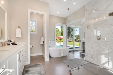 Sunlit modern master bathroom with freestanding white tub, glass-enclosed marble shower, double vanity, and sliding doors opening to a backyard pool and patio umbrella.