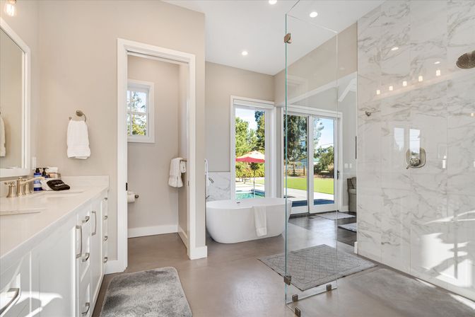 Sunlit modern master bathroom with freestanding white tub, glass-enclosed marble shower, double vanity, and sliding doors opening to a backyard pool and patio umbrella.