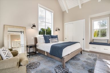Sunlit modern farmhouse bedroom with vaulted white-beamed ceiling, large window overlooking greenery, wooden platform bed with white linens and navy throw, built-in window seat with cushions, patterned blue area rug, leaning mirror, armchair and wall sconces.