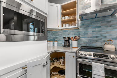 Bright modern kitchen corner with white cabinets and blue subway tile backsplash, stainless steel microwave and range, open cabinets showing glassware and pots, and countertop coffee maker and kettle.