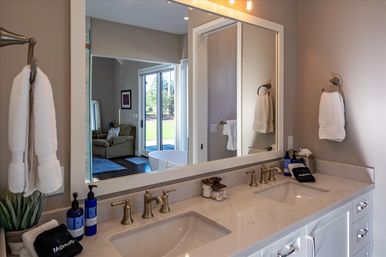 Spacious modern bathroom with double-sink marble vanity, brass faucets, large mirror reflecting a freestanding tub and sliding glass doors to a sunny backyard, with white towels and neatly arranged toiletries.