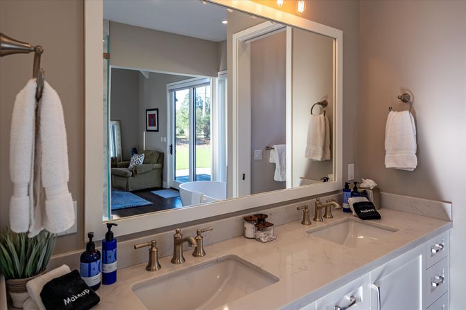 Spacious modern bathroom with double-sink marble vanity, brass faucets, large mirror reflecting a freestanding tub and sliding glass doors to a sunny backyard, with white towels and neatly arranged toiletries.