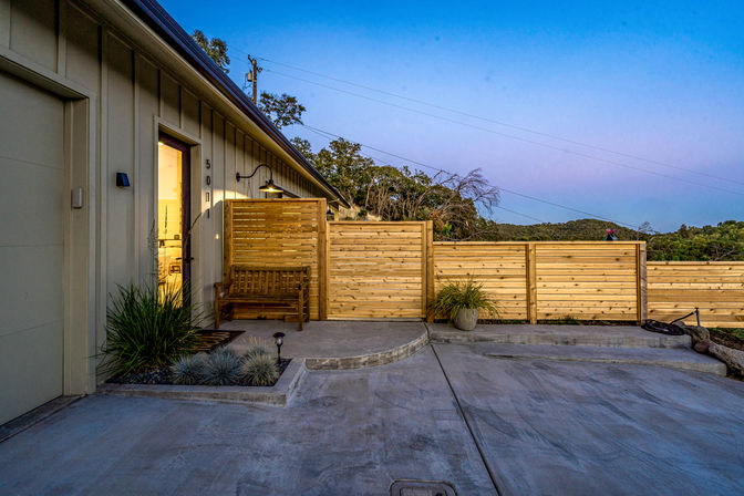 Modern home exterior at dusk with wooden privacy fence, bench, potted plants and concrete driveway overlooking rolling wooded hills