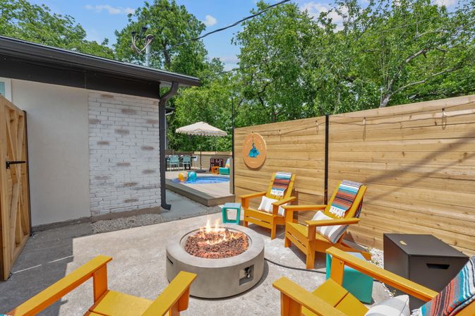 Sunny backyard patio with round concrete fire pit surrounded by four bright orange Adirondack chairs draped with colorful striped throws, wood privacy fence, and a small decked plunge pool with umbrella and inflatables in the background.