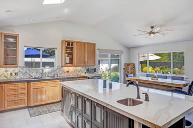 Bright open-concept kitchen with large marble island and sink, dark island base, wood cabinets, herringbone tile backsplash, dining table by sliding glass doors opening to a sunny backyard patio with umbrella