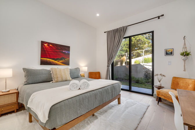 Bright modern bedroom with a large bed in gray linens, rolled towels, wooden nightstands and lamps, sliding glass doors to a sunny garden patio, orange accent chairs and colorful wall art.