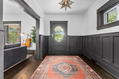 Welcoming entryway with dark gray wainscoting and oval-glass front door, hardwood floors, a patterned red Persian-style rug, star-shaped pendant light, and a glimpse of a gray living room and greenery outside.