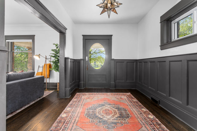 Welcoming entryway with dark gray wainscoting and oval-glass front door, hardwood floors, a patterned red Persian-style rug, star-shaped pendant light, and a glimpse of a gray living room and greenery outside.