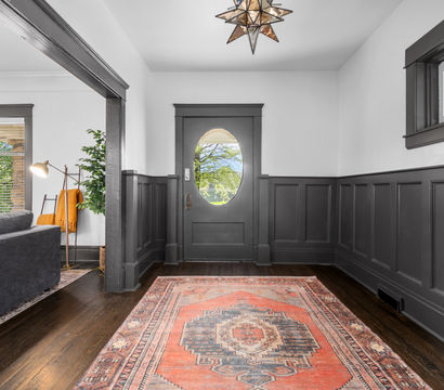 Welcoming entryway with dark gray wainscoting and oval-glass front door, hardwood floors, a patterned red Persian-style rug, star-shaped pendant light, and a glimpse of a gray living room and greenery outside.