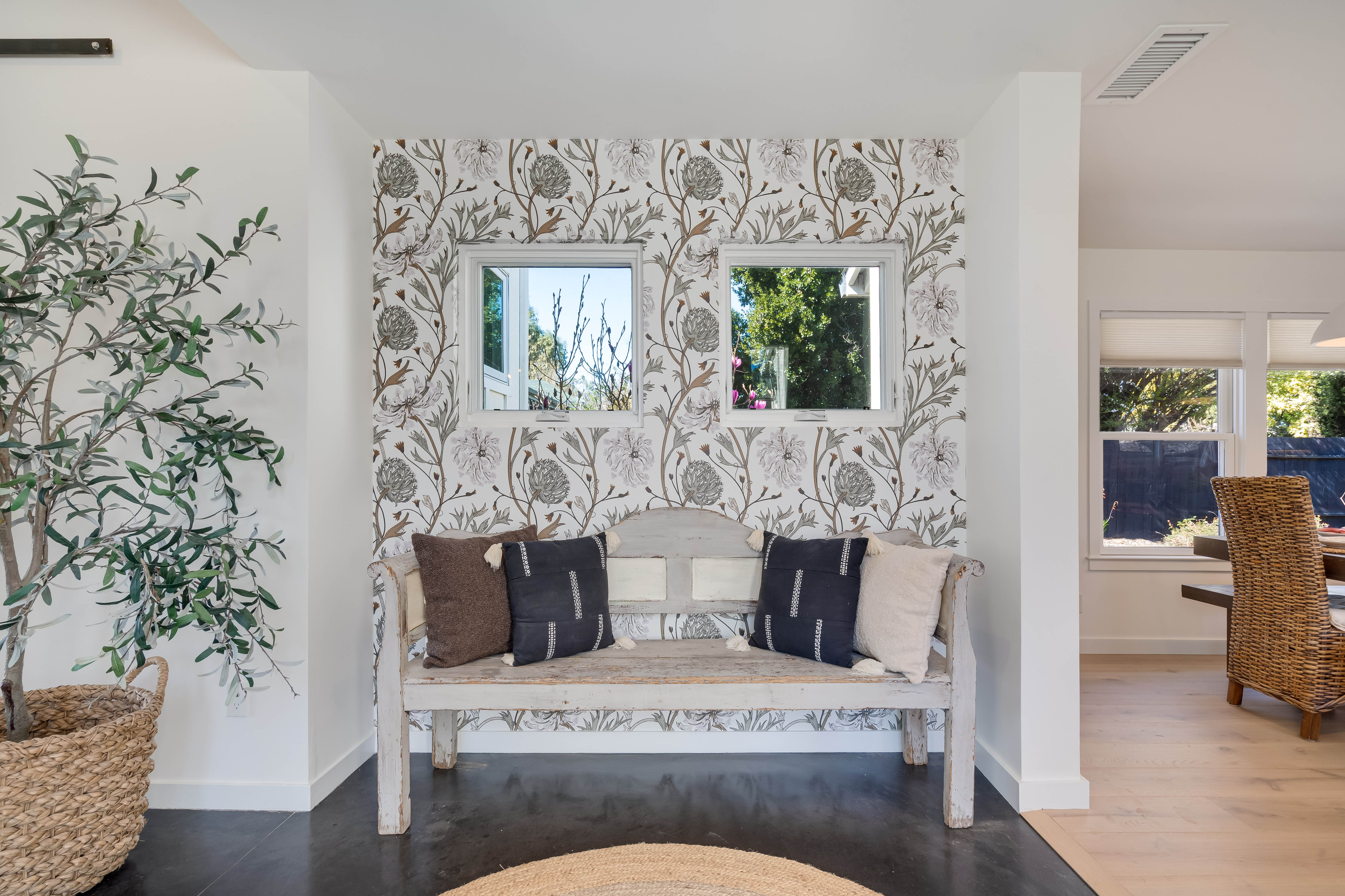 Cozy modern farmhouse entryway: distressed white wooden bench with mixed throw pillows against floral wallpaper and two small windows, a potted olive tree in a woven basket, dining area glimpsed to the right.