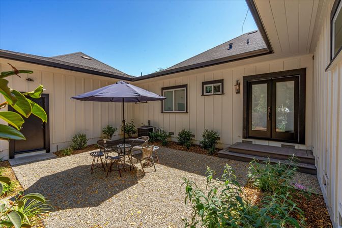 Sunny private residential courtyard patio with gravel floor, black bistro table and chairs under a navy umbrella, white board-and-batten siding, double French doors and small shrubs and potted plants