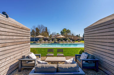 Inviting poolside retreat framed by wooden privacy walls, cozy outdoor lounge with cushioned sofas and table, green lawn leading to an in-ground swimming pool with sun loungers and umbrellas under a clear blue sky.