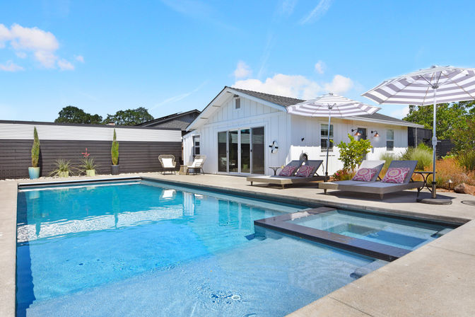 Sunny modern backyard with rectangular swimming pool and built-in spa beside a white ranch-style home, patio loungers with patterned cushions and striped umbrellas.
