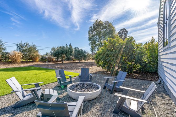 Sunny suburban backyard patio with circular stone fire pit surrounded by six dark gray Adirondack chairs on gravel, adjacent bright green lawn, mature trees and blue sky with sun rays.