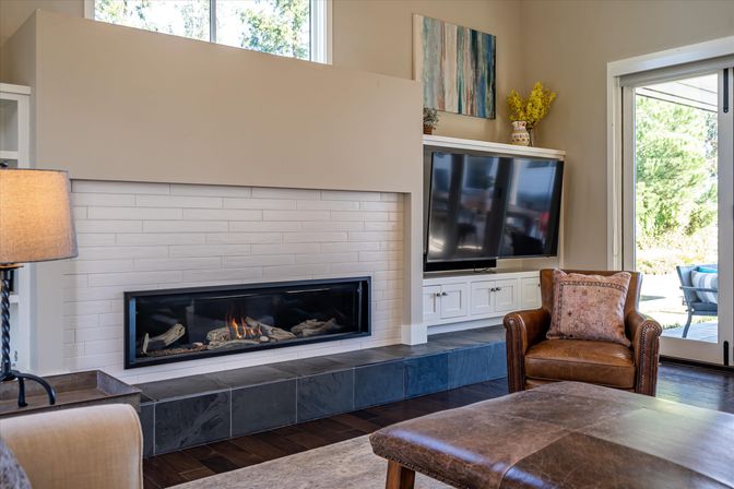 Cozy modern living room with linear gas fireplace, wall-mounted flat-screen TV, leather armchair and sliding glass door to a sunny patio