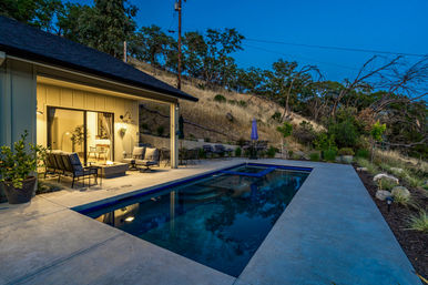 Twilight backyard pool scene: rectangular plunge pool with concrete deck, cozy patio seating and fire pit outside a lit modern home, set against a dry grassy hillside.