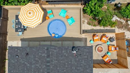 Aerial view of a residential backyard deck and patio with a yellow-striped umbrella over a dining table, a round blue plunge pool, turquoise lounge chairs, and orange armchairs gathered around a fire pit.