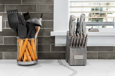 Modern kitchen countertop with wooden-handled utensils in a metal holder and a stainless-steel knife block against gray subway tile backsplash and window blinds