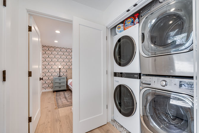 Stacked front‑load washer and dryer in a compact laundry closet with detergent on the shelf, open white door revealing a bedroom with patterned accent wall, nightstand, lamp and light hardwood floors.