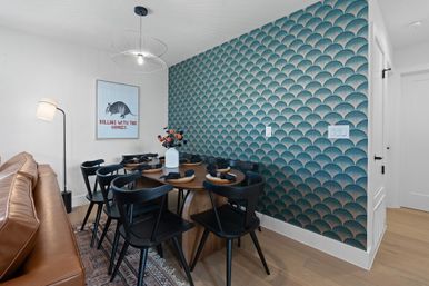 Modern dining nook in an apartment with teal scallop-pattern accent wall, round wood table set for six with black chairs, tan leather sofa, wire pendant light, floor lamp, and a playful armadillo poster.