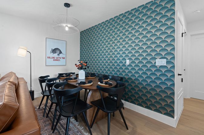Modern dining nook in an apartment with teal scallop-pattern accent wall, round wood table set for six with black chairs, tan leather sofa, wire pendant light, floor lamp, and a playful armadillo poster.