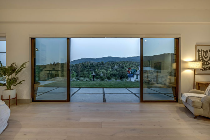 Modern living room with wide sliding glass doors opening to a patio and lush hillside/mountain views at dusk, light hardwood floors, potted plant, cozy armchair and lamp.