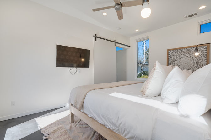 Bright modern bedroom with white linens on a wooden bed, decorative mandala headboard, wall-mounted TV, barn-style sliding door, ceiling fan, and sunlight through a window.