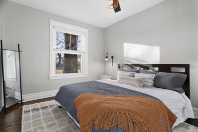 Cozy, light-filled bedroom with gray walls, dark wood headboard, navy bedding, striped pillows and a rust-colored throw on a patterned rug, large window showing a tree outside.