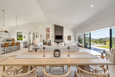 Bright modern open-plan hilltop living space with a long wooden dining table and curved chairs, neutral sofas around a dark stone fireplace, kitchen island with leather barstools and pendant lights, and floor-to-ceiling sliding glass doors opening to a pool and sunny hillside view.