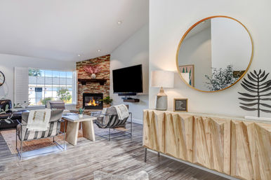 Sunlit modern suburban living room with vaulted ceiling, stone fireplace and wall-mounted TV, striped accent chairs around a coffee table, large window, and geometric wood console topped by a round mirror and lamp.