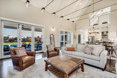 Sunlit modern farmhouse open-concept living room with white beamed vaulted ceiling, leather club chairs, neutral sofa and large leather ottoman, sliding glass doors to patio and backyard pool with red umbrellas, and a kitchen island with chandelier in the background.