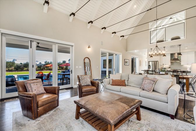 Sunlit modern farmhouse open-concept living room with white beamed vaulted ceiling, leather club chairs, neutral sofa and large leather ottoman, sliding glass doors to patio and backyard pool with red umbrellas, and a kitchen island with chandelier in the background.