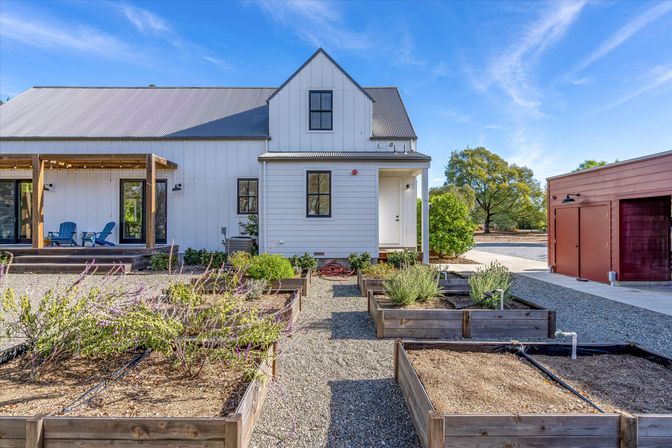 Modern white farmhouse with covered porch and blue Adirondack chairs, multiple raised wooden garden beds and gravel paths, red detached garage and oak trees under a bright blue sky.