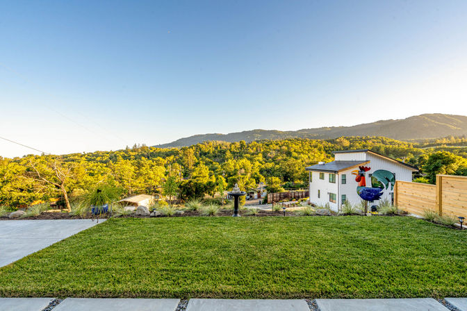 Hillside backyard with landscaped lawn and concrete patio, decorative metal rooster sculpture and birdbath, overlooking a sunlit forested valley and distant mountains under a clear blue sky.