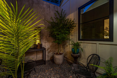 Cozy leafy urban patio at night with pebble floor, potted palm and small tree, teal planter on a pedestal, black wire chairs and warm accent lighting by a window.