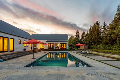 Modern farmhouse backyard with rectangular pool at sunset, red patio umbrellas and lounge chairs on a concrete deck, warm interior lights reflecting in the water, and tall trees framing a landscaped yard.