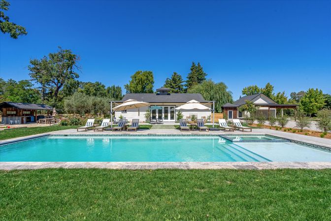 Sun-soaked rectangular backyard pool with clear turquoise water, stone deck lined with lounge chairs and umbrellas, a white farmhouse-style house, manicured lawn and trees under a bright blue sky.