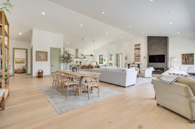 Bright open-concept modern living room and kitchen with light hardwood floors, wooden dining table, kitchen island with pendant lights, gray sofas, and a concrete fireplace with wall-mounted TV.