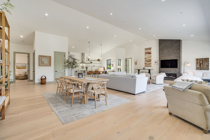 Bright open-concept modern living room and kitchen with light hardwood floors, wooden dining table, kitchen island with pendant lights, gray sofas, and a concrete fireplace with wall-mounted TV.
