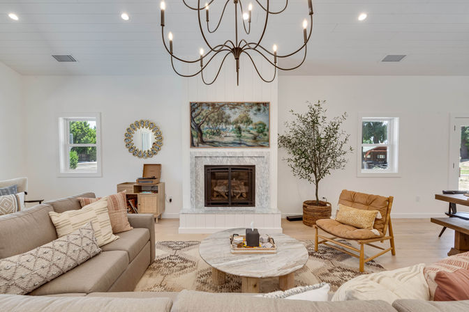 Bright cozy modern farmhouse living room with marble fireplace, oversized chandelier, neutral sectional, round marble coffee table, woven rug, and potted olive tree
