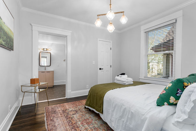 Bright bedroom with hardwood floors, white walls and crown molding, queen bed topped with a green throw and playful butterfly pillow, folded towels on the foot of the bed, patterned area rug, brass modern chandelier, glass side table, and an open doorway revealing a small bathroom vanity and mirror.
