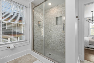 Bright, spa-like home bathroom with a glass-enclosed walk-in shower, neutral geometric wall tiles and mosaic niche, sliding glass door, white window blinds and beige floor tile.