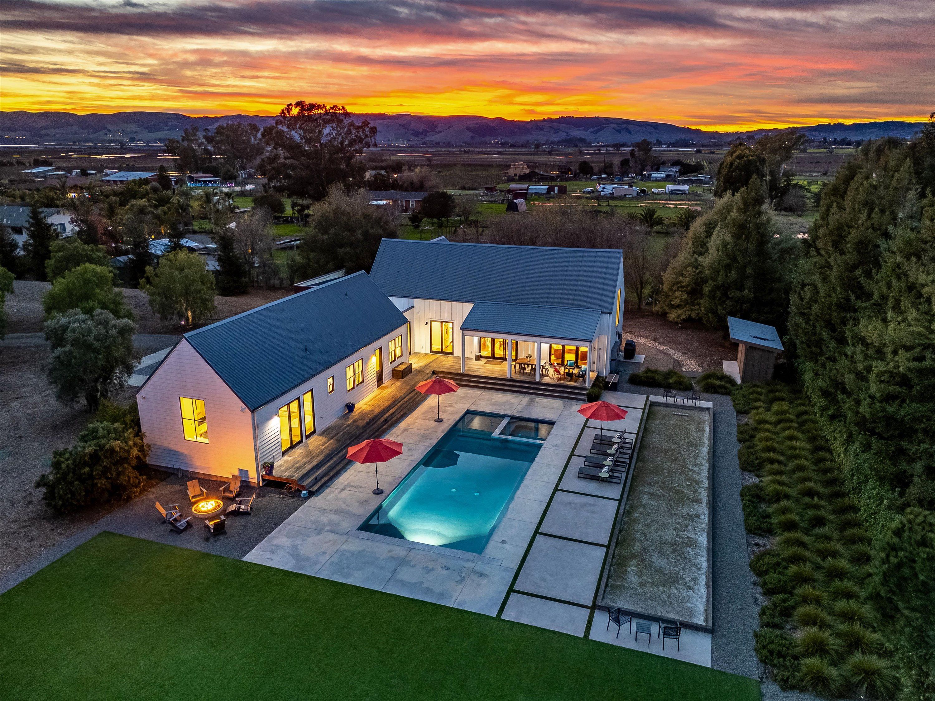 Aerial dusk view of a modern farmhouse in the countryside with glowing windows, a concrete patio and rectangular swimming pool with red umbrellas, lounge chairs and a fire pit, surrounded by fields and rolling hills at sunset.